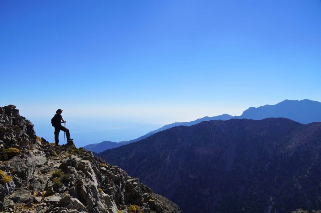 A panoramic view from a Greek mountain peak looking over ridges toward the Mediterranean Sea during ideal hiking conditions.