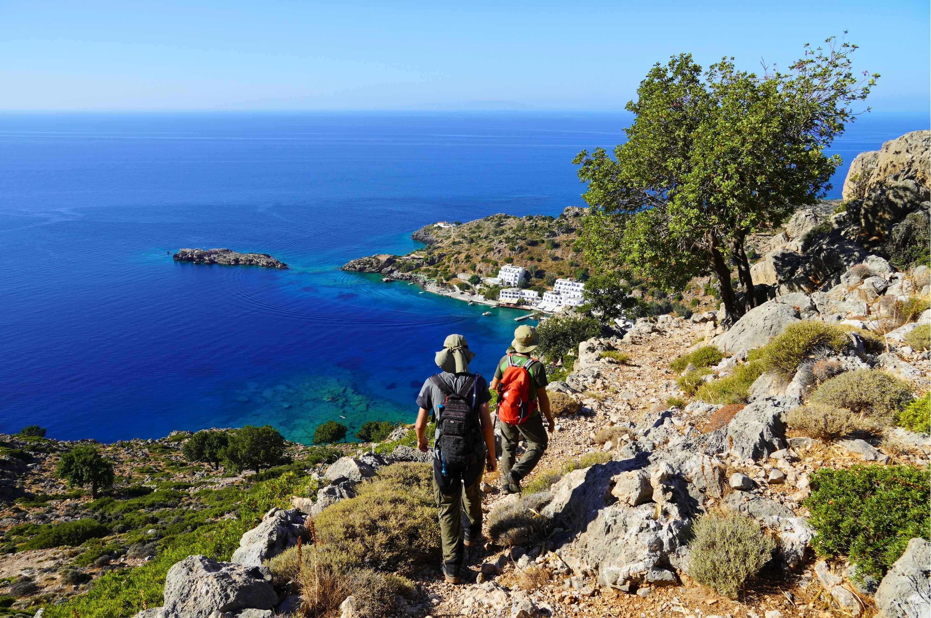 Two hikers descending a rocky trail overlooking a deep blue turquoise bay and a traditional white coastal village in Crete, Greece.