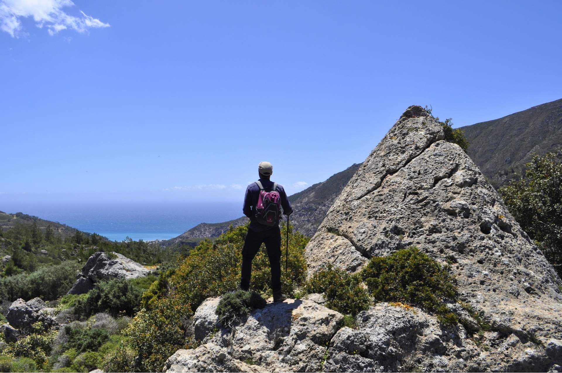 A hiker with a backpack stands on a rocky outcrop looking down at a traditional coastal village and the blue sea in Southwestern Crete.