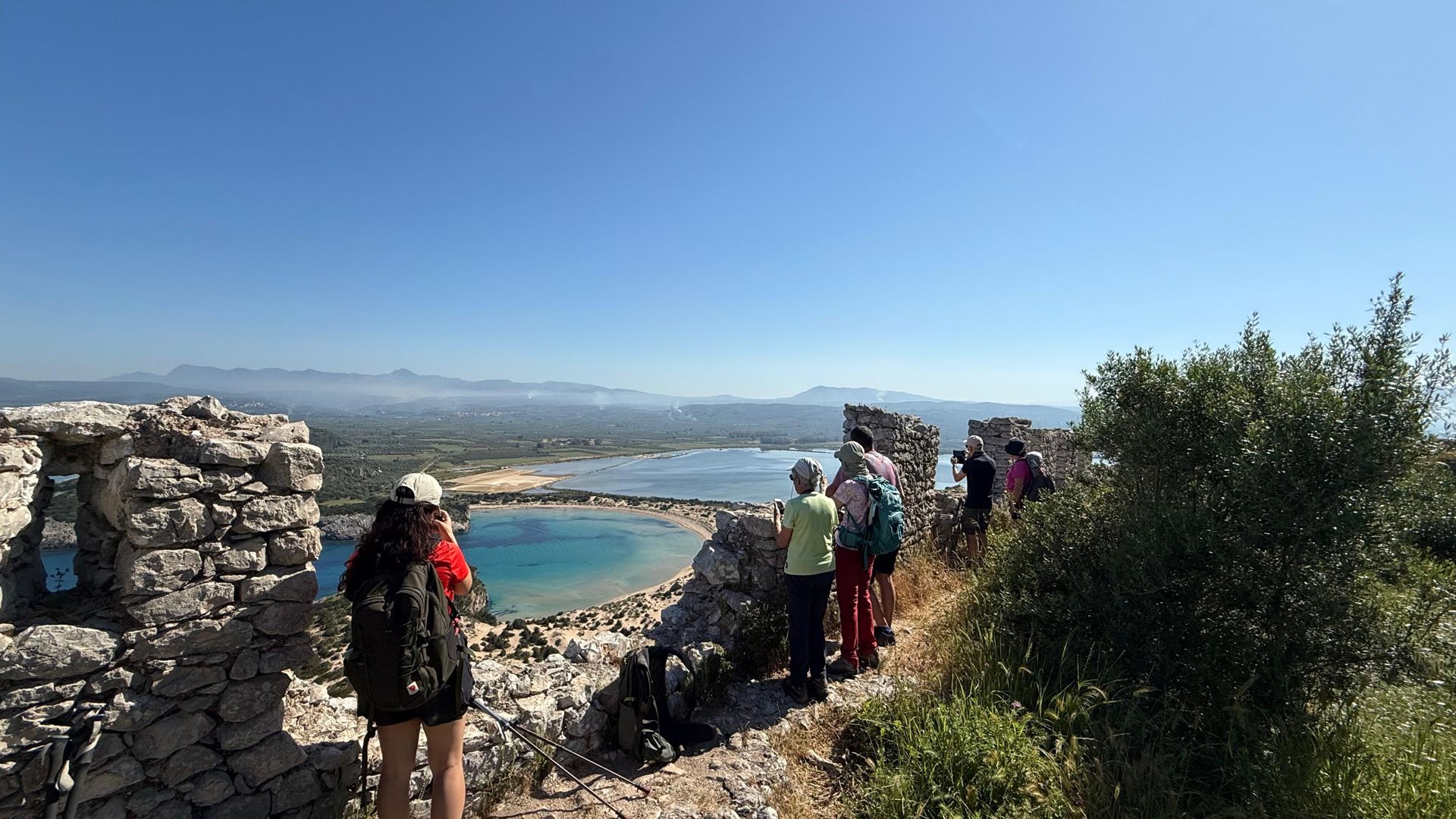 Hikers looking down on a turquoise coastal bay from the ruins of a hilltop castle in Messenia, Peloponnese